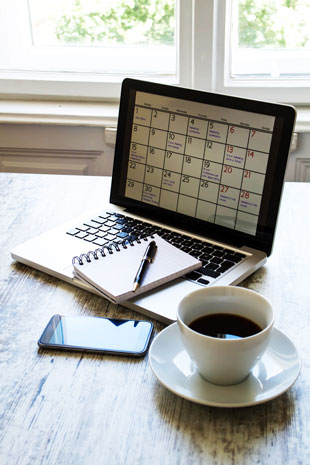 geöffnetes Notebook, Kalenderanzeige im Display mit Notizblock, Kugelschreiber und Kaffeetasse