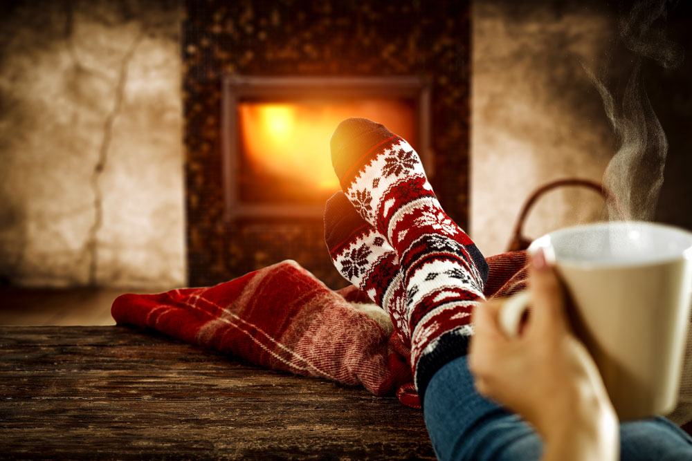 Weihnachtszeit: Frau mit Wintersocken, die eine Tasse in der Hand hält und vor einem brennenden Kamin sitzt