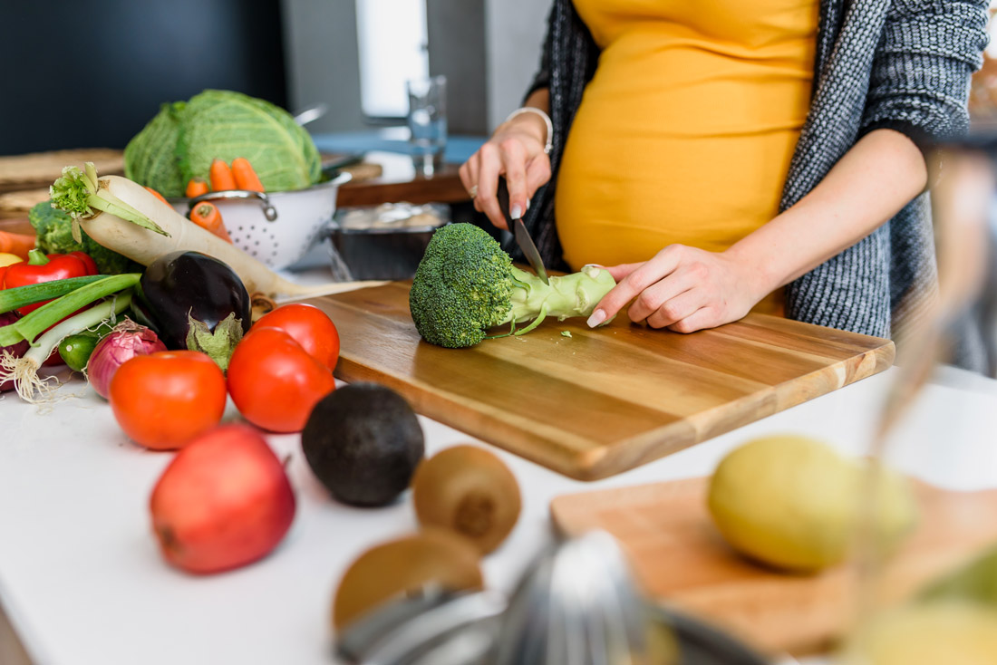 schwangerschaft Eine schwangere Frau steht in der Küche und schneidet Obst und Gemüse auf einem Holzbrett.