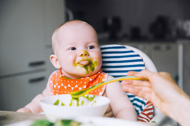Ein im Hochstuhl sitzendes Baby wird mit grünem Püree gefüttert