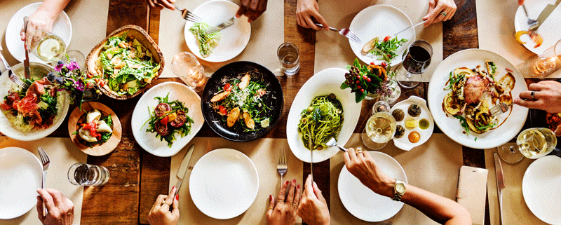 Eine Tischtafel, es stehen Gerichte in der Mitte, die Personen bedienen sich daran.