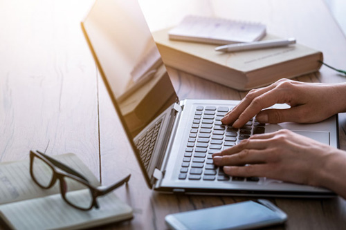 Eine Person sitzt an einem Laptop. Auf dem Tisch liegt ein Buch, Notizbücher, Smartphone, Brille, ein Stift.