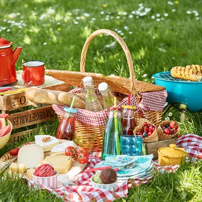 Auf grüner Wiese gefüllter Picknickkorb, Grill mit Maiskolben, Käseauswahl, Baguette, Melone, Eis, Früchte, dazu Kaffee und Limonate